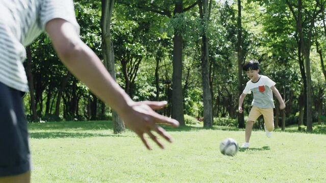 Asian father playing soccer with son on park lawn