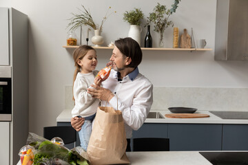 Happy dad holding adorable little daughter child in arms, unpacking grocery paper bag in kitchen, talking to kid, smiling, laughing, enjoying leisure, family household chores together
