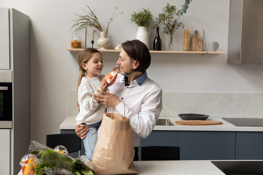 Happy Dad Holding Adorable Little Daughter Child In Arms, Unpacking Grocery Paper Bag In Kitchen, Talking To Kid, Smiling, Laughing, Enjoying Leisure, Family Household Chores Together