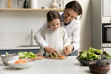 Happy joyful daddy teaching little daughter kid to cook salad, slicing fresh vegetables on chopping...