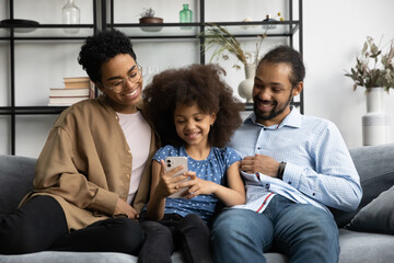 Happy Afro American family using online app on mobile phone, having fun at home. Parents, mom, dad,...