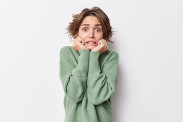 Worried stressed young woman keeps hands under chin afraids of something wears casual jumper stands embarrassed poses against white background reacts to scary news poses indoor. Human reaction