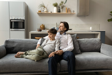 Happy father and teen son resting on couch. Dad spending leisure time with teenage kid, sitting on sofa, talking to boy, chatting, laughing, hugging child, looking at window away. Fatherhood concept