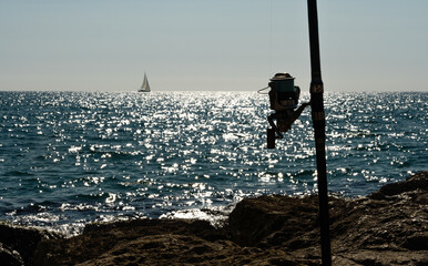 Close-up backlight of fishing rod with sea and sailboat in the background, selective focus.