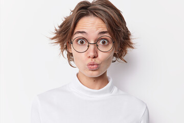 Fototapeta premium Portrait of surprised dark haired woman pouts lips as if wanting to kiss someone wears round spectacles for vision correction casual jumper isolated over white background. Human face expressions