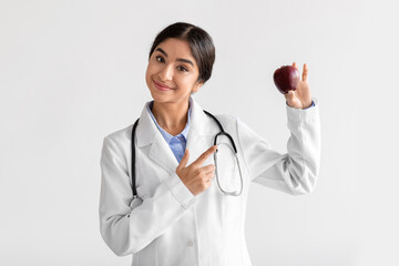 Smiling friendly millennial cute indian lady doctor in uniform shows finger on red apple