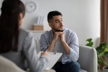 Depressed arab man consulting psychologist, sitting on couch deep in thought and looking aside, having session in clinic