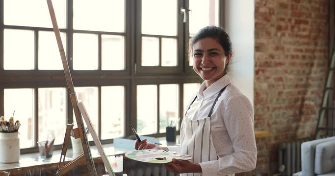 Portrait happy smiling young Indian lady artist amateur stand near easel with paintbrush in hand proud with nice picture. Excited woman painter enjoy working with oil on canvas at school home studio