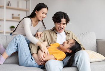 Happy teenager girl and millennial asian man and woman have fun together, tickling little daughter on sofa - obrazy, fototapety, plakaty