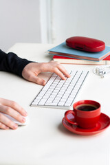 Man hands in lay on keyboard and mouse with red cup of coffee, notebooks, headhones and glasses case on white table - 479336552