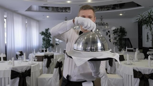 A cheerful male waiter is having fun in a restaurant, he puts a metal lid on his head while standing in a restaurant.