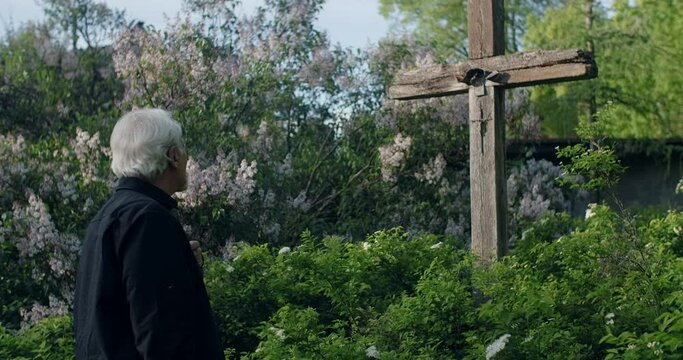 Sad old gray haired man stands near big old wooden cross at the cemetery. Sorrow and sadness about lost of close people. Religious man, 4k 60p Prores
