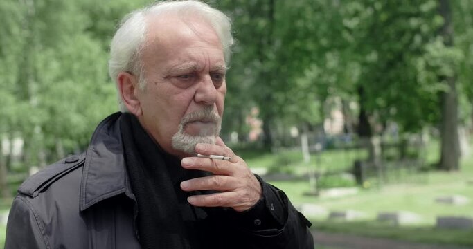 Sad old gray haired man smokes cigarette on the cemetery, sorrow and sadness about lost of close people, 4k 60p Prores