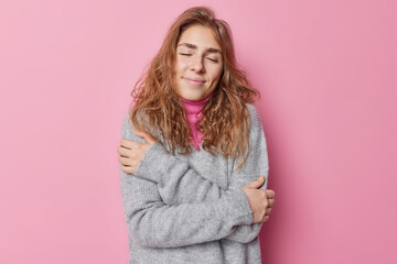 Love yourself. Pretty long haired young woman embraces herself enjoys softness of jumper has dreamy expression stands with closed eyes isolated over pink background. Self acceptance concept.