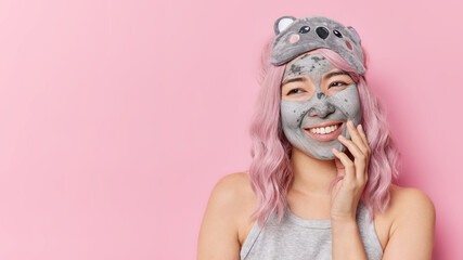 Happy Asian woman with dyed hair applies clay mask on face for softness of skin wears sleepmask casual t shirt looks gladfully away isolated over pink background blank copy space for your text