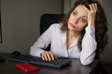 Tired young woman with curly hair and white shirt is working at the office using her laptop, routine work, freelance, burnout syndrome