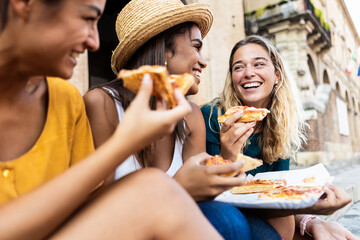 Three young multiracial women laughing while eating a piece of pizza in italian city street - Happy...