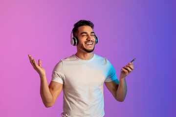 Cool Arab man listening to popular song in headphones, checking new mobile music app on phone,...