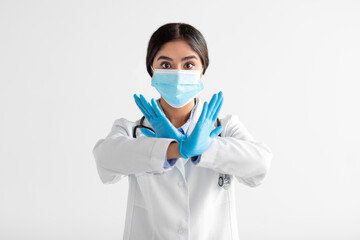 Serious young indian lady doctor in uniform, gloves and protective mask show sign with hands, say no, cross arms