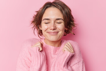 Pleased beautiful woman shakes hands keeps eyes closed awaits for surprise smiles joyfully wears soft jumper isolated over pink background. Cheerful brunette female anticipates for smth pleasant