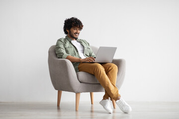 Happy indian guy in casual sitting in armchair with laptop