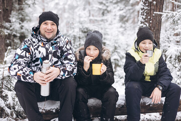 Father and sons drinking tea from thermos and talking sitting together on log in winter snowy...