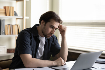 Thoughtful unhappy young male employee looking at laptop screen, feeling stressed stack with difficult task, getting email with negative news or dismissal notice, thinking of financial problems.