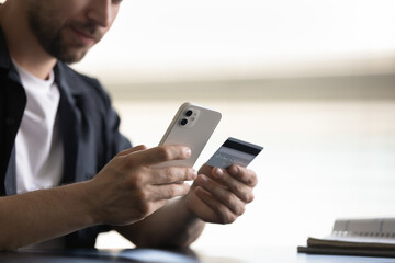 Close up cropped happy focused young man holding plastic bank card and cellphone in hands, making payments in app, purchasing goods or service online, transferring money, financial operation security.