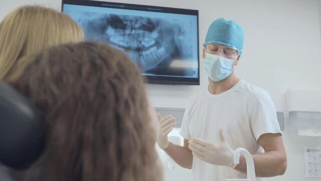 Middle shot of a male Caucasian dentist with protective gloves, face mask, and surgical cap advising and showing to a patient, a female child, and her mother an X-ray of the teeth on the screen.