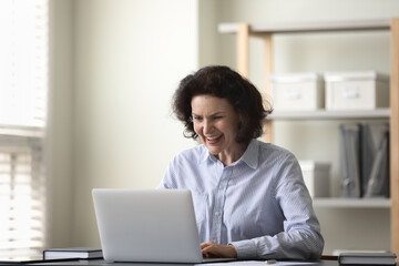 Fototapeta premium Joyful middle aged 60s woman looking at laptop screen, feeling excited reading email with amazing news, getting bank loan approval or money refund, celebrating business achievement at home office.