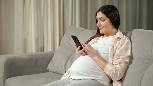 Young pregnant woman with dark long hair in comfortable outfit scrolls through social media using smartphone and sitting on sofa in living room.