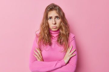 Fototapeta premium Upset offended young woman has gloomy sullen expression holds hands crossed wears casual turtleneck feels sad or hurt waits for apologizing poses against pink background. Candid emotions concept