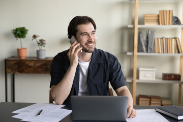 Naklejka premium Happy friendly millennial employee businessman multitasking in modern workplace, communicating distantly by phone call with client, discussing online project, working on computer, looking in distance.