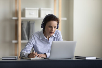 Happy concentrated middle aged elderly woman in wireless headphones looking at computer screen, studying on online courses, improving knowledge, e-learning using video call application, writing notes.