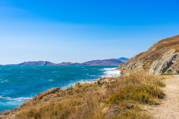 Obraz premium Picturesque summer landscape of sea coast with waves on water surface against mountains and sky. Beautiful seascape in sunny day, natural background. Feodosia vicinity, Crimea, Russia
