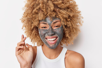 Beauty portrait of happy curly haired woman applies moisturizing mask on face smiles broadly shows white teeth wears casual t shirt poses indoor. Wellness skin care and rejuvenation concept.