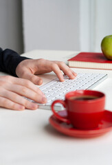 Man hands on wireless keyboard with red notebook, green apple and red cup of coffee on white table - 479354332