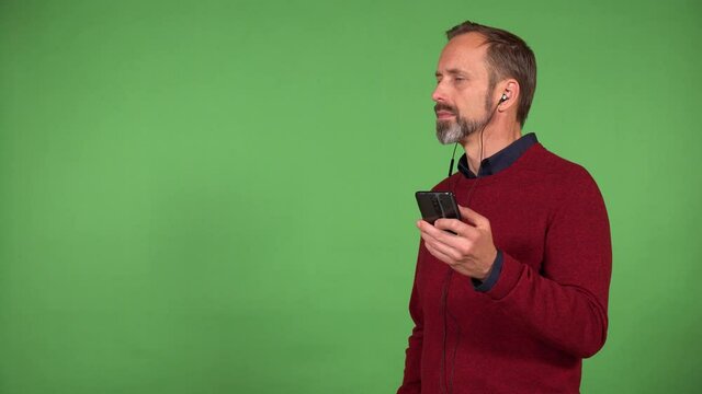 A middle-aged handsome Caucasian man listens to something with earphones on and a smartphone - green screen background