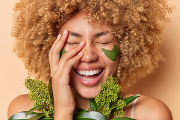 Beauty and skin care concept. Overjoyed woman laughs out happily covers face with hand smiles toothily has natural clean skin applies green collagen patches under eyes for reducing wrinkles. - obrazy, fototapety, plakaty