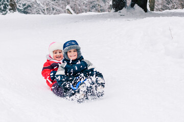Little caucasian kids in bright warm clothing laughing looking at camera and toboganning on snow covered hill. Full lengh horizontal shot. Happy childhood and active wintertime concept.