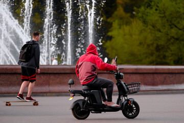A man is riding a scooter in the park, a teenager is riding a skateboard next to a fountain. People are actively relaxing in the city park.