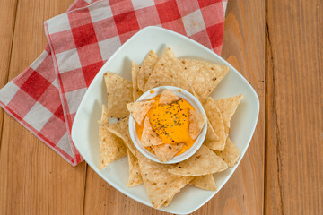 Nachos dipped in cheddar on a bowl on a wooden table. Under the dish there is a red and white kitchen cloth and the background is grey. Vertical image