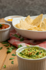 Bowls with red pepper sauce, cheddar and guacamole surrounding a dish with nachos on a wooden table. On the table there also are a red and white kitchen cloth and coriander leaves. Vertical image