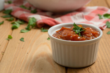 Bowl with a red pepper sauce and a dish with nachos in the back on a wooden table. On the table there also are a red and white kitchen cloth and coriander leaves. Horizontal image