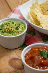 Bowls with red pepper sauce and guacamole, and a dish with nachos on a wooden table. On the table there also are a red and white kitchen cloth and coriander leaves. Vertical image