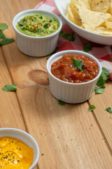 Bowls with red pepper sauce, cheddar and guacamole, and a dish with nachos in the back on a wooden table. On the table there also are a red and white kitchen cloth and coriander leaves. Vertical image