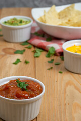 Bowls with red pepper sauce, cheddar and guacamole, and a dish with nachos in the back on a wooden table. On the table there also are a red and white kitchen cloth and coriander leaves. Vertical image