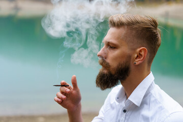 Concept. Smoke enveloped man head against backdrop of reservoir in park. Portrait of a bearded...