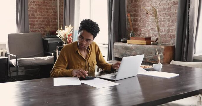 Serious busy African female accountant sit at desk with laptop, do accountancy job calculates summary to pay make online payment via e-bank application. Secure software, finances management concept