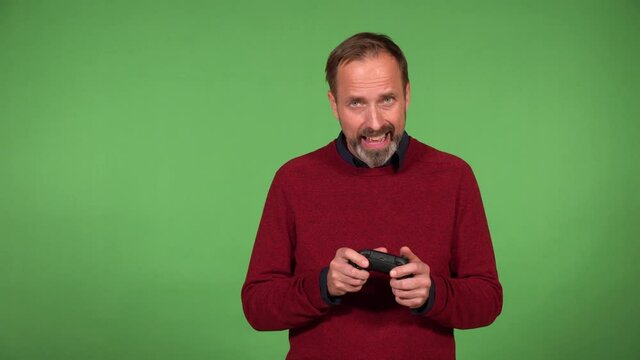 A middle-aged handsome Caucasian man plays a game on the camera and holds a controller - green screen background